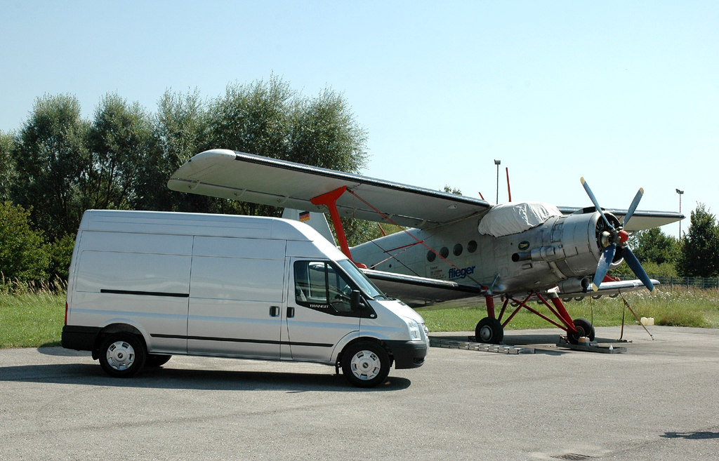 Ford Transit, hier als Kastenwagen mit Hochdach, vor einem Antonow-Doppeldecker vor der (Museums-)Flugwerft in Oberschleißheim. 